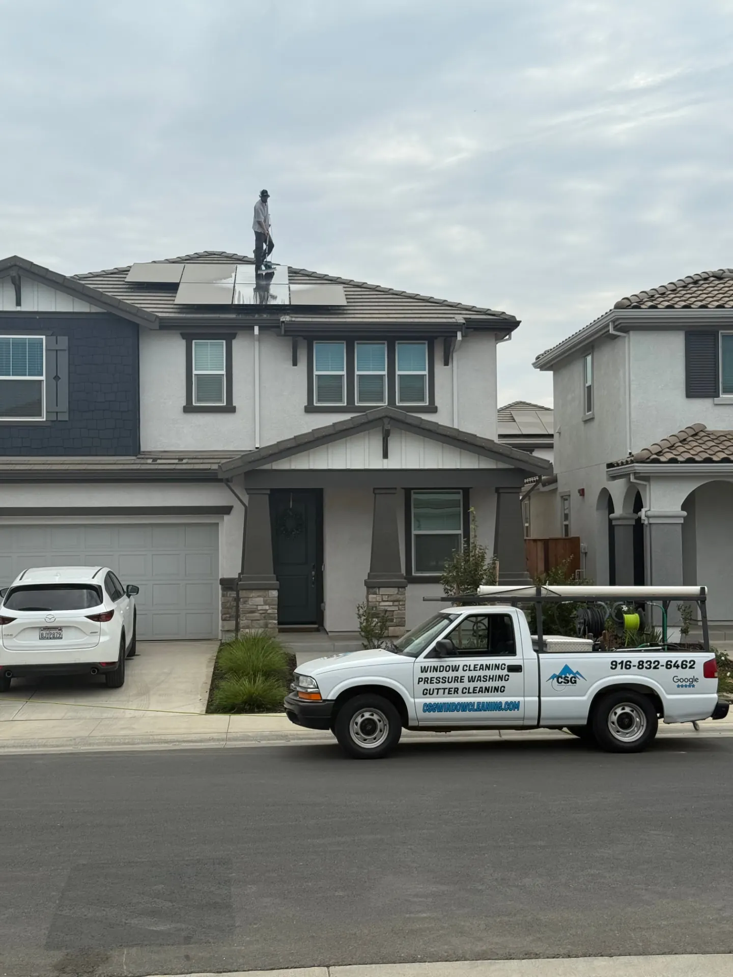 CSG technician rinsing a rooftop solar array on a two-story Elk Grove home with the CSG service truck parked out front